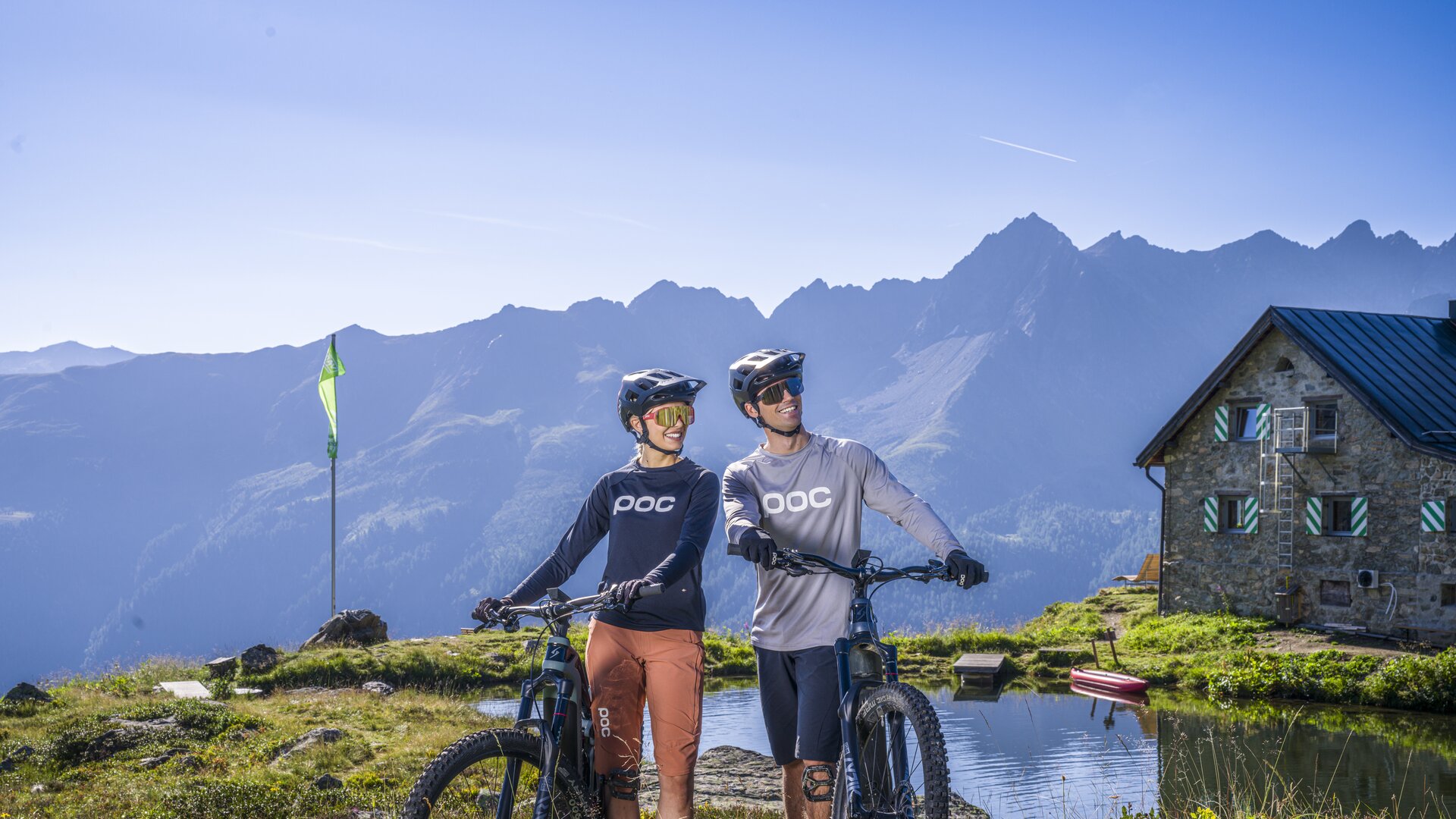 Ein Mann und eine Frau in schicken Mountainbike-Outfits stehen in den Ischgler Bergen hinter einer Hütte neben einem glitzernden See und lächeln fröhlich.