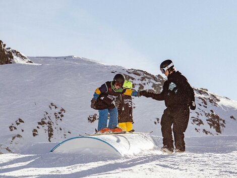 Ein Junge übt das Snowboardfahren auf einem Schneehügel. Ein Trainer unterstützt ihn dabei in einer winterlichen Landschaft.