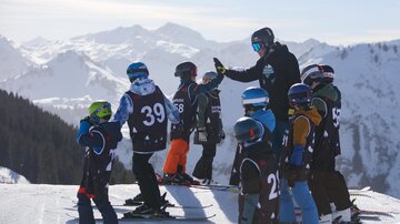Eine Gruppe von Kindern steht auf Skiern und schaut zu einem Lehrer, der ihnen Anweisungen gibt. Im Hintergrund sind schneebedeckte Berge und ein klarer Himmel zu sehen.