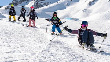 Eine Gruppe von Kindern fährt Ski in einem verschneiten Gebirge. Sie tragen helle Skikleidung und genießen den Tag auf der Piste.