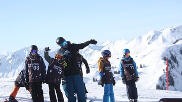 Eine Gruppe von Skifahrern und Snowboardern steht auf einem schneebedeckten Berg. Im Hintergrund sind majestätische Berge und blauer Himmel zu sehen.