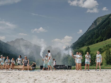 A group of people is standing on a sandy shore amidst mountains. Some sit on benches, while others seem to be participating in a game or an activity.