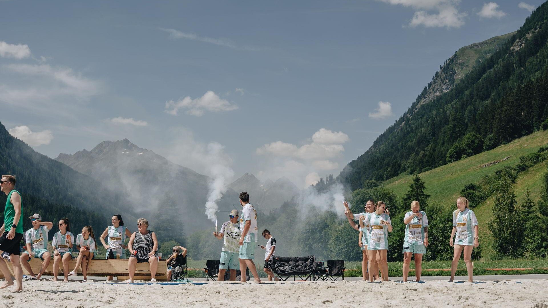 A group of people is standing on a sandy shore amidst mountains. Some sit on benches, while others seem to be participating in a game or an activity.