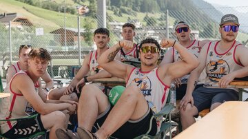 A group of young men is sitting relaxed at a table outside. They are wearing sporty shirts and sunglasses and seem to be having fun.