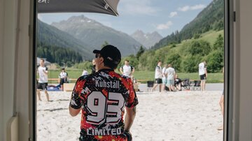 A man sits in the sand and looks at a group of people playing actively. In the background, mountains and green countryside can be seen.