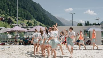 A group of young people is playing in the sand and having fun. In the background, mountains and sunshades can be seen.