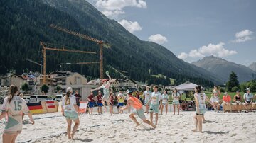 A group of young athletes is playing beach volleyball on the beach. In the background, mountains and a busy village can be seen.
