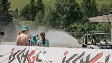 A group of people is standing at a venue while water is being sprayed into the air. In the background, green trees and a landscape can be seen.