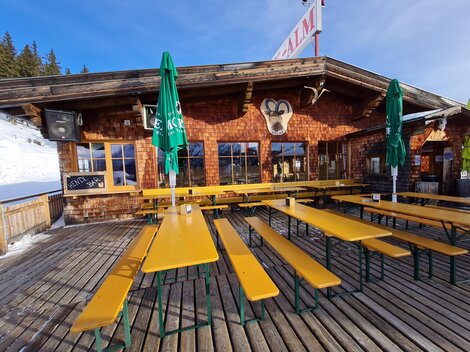 A rustic mountain cabin with a wooden terrace and yellow benches. There are green umbrellas and a snowy landscape in the background.
