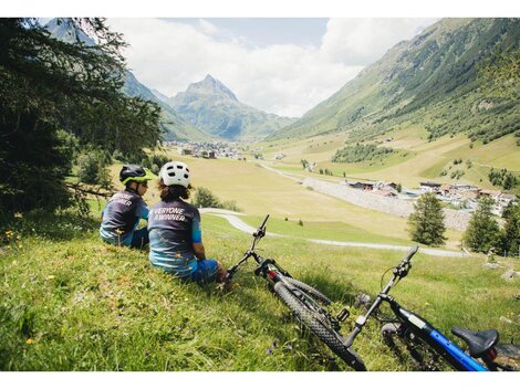 Two children are sitting on a meadow and looking at the valley with a village in the background. Next to them are two bicycles, surrounded by a beautiful mountain landscape.