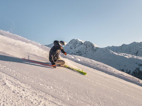 Ein Skifahrer fährt die schneebedeckte Piste hinunter. Im Hintergrund sind majestätische Berge zu sehen.