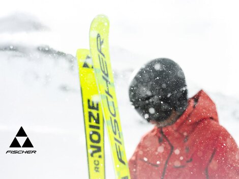 A skier in a red suit stands in the snow. The surroundings are wintry and snowy, while the skis are bright yellow.