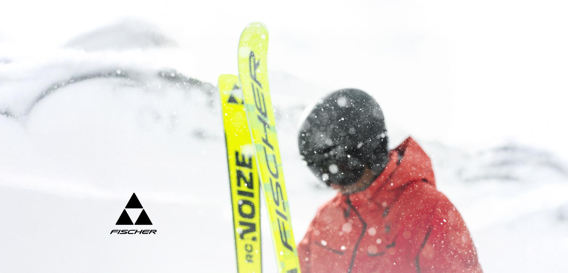 A skier in a red suit stands in the snow. The surroundings are wintry and snowy, while the skis are bright yellow.