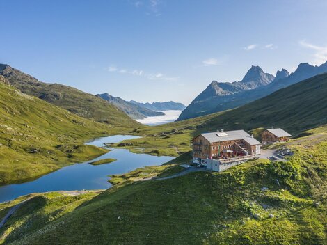Ein modernes Holzgebäude liegt malerisch in den Bergen. Im Hintergrund sind schneebedeckte Gipfel und eine grüne Landschaft zu sehen.