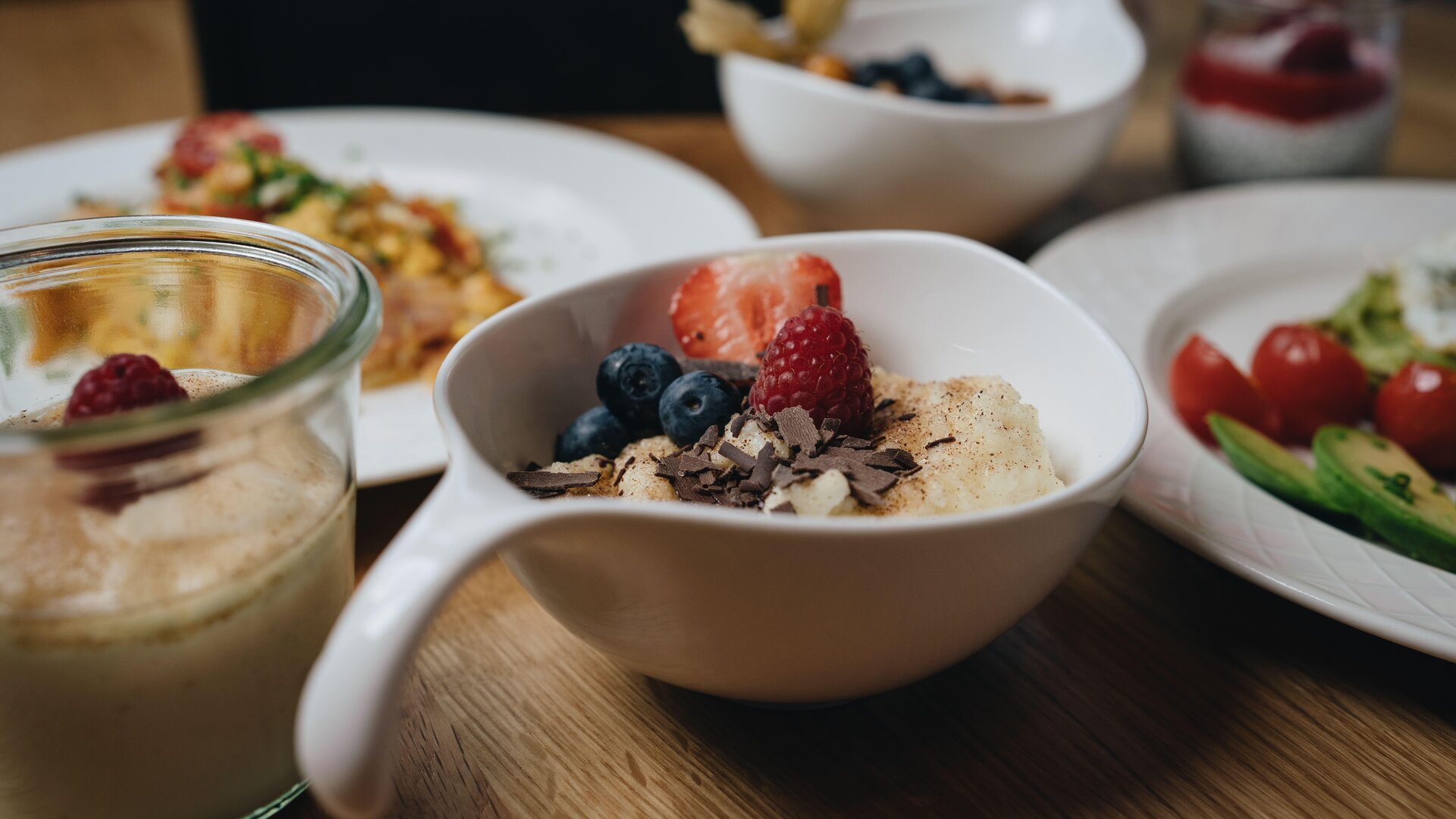 Ein schöner Tisch mit verschiedenen Frühstücksgerichten. Eine Schüssel mit Haferbrei, frischen Beeren und Schokolade ist im Vordergrund zu sehen.