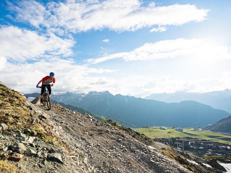 A mountain biker rides over a steep, rocky path with a spectacular mountain landscape in the background. The sky is clear and there are many clouds.