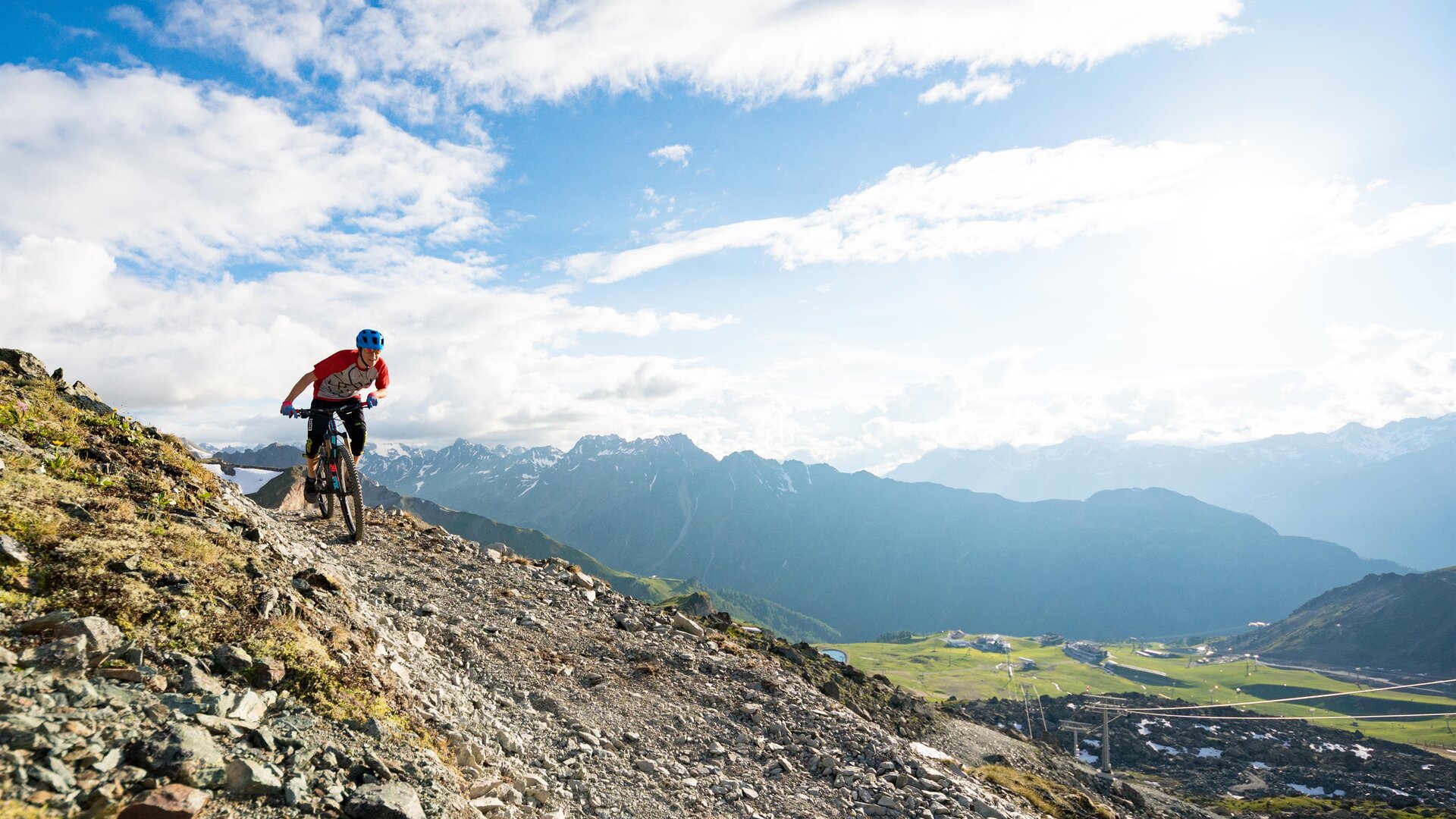 A mountain biker rides over a steep, rocky path with a spectacular mountain landscape in the background. The sky is clear and there are many clouds.
