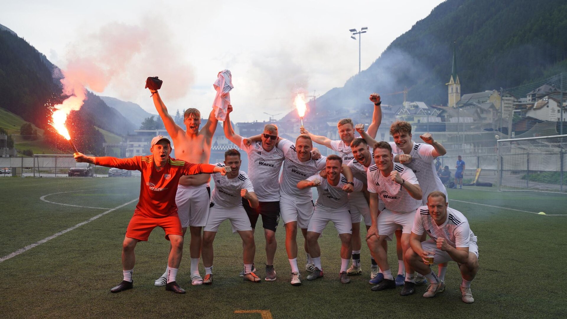 A group of soccer players is celebrating on the field. They are cheerful and holding torches in their hands, while a picturesque mountain landscape can be seen in the background.