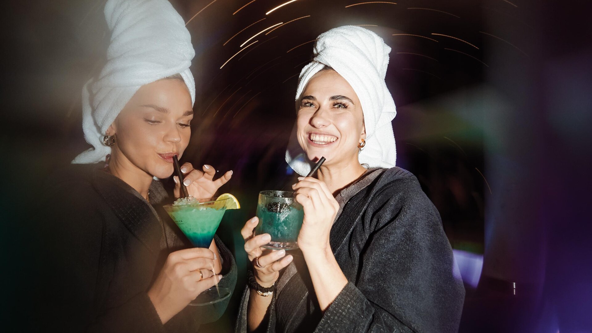 Two women in bathrobes and with towels on their heads are holding colorful cocktails. In the background, lights and a relaxed atmosphere can be seen.