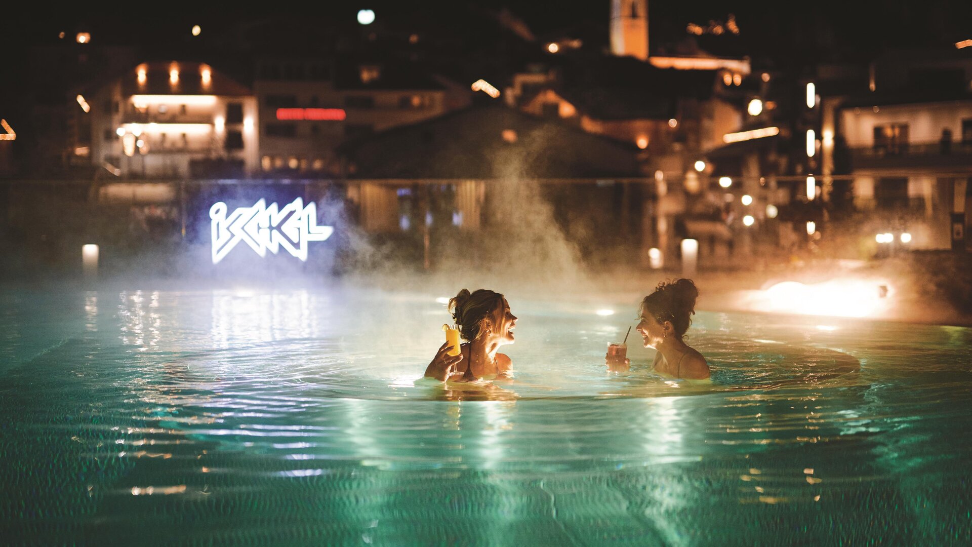 Zwei Frauen entspannen im beleuchteten Pool bei Nacht. Im Hintergrund sind die Lichter der Stadt sichtbar.