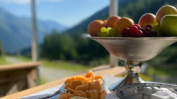 A beautiful view of the mountains with fresh fruit on a table. In the foreground, cut fruits and a bowl with more fruit can be seen.