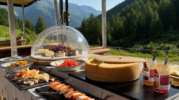 A festively arranged table outdoors with an assortment of cheese, sausage, and fresh vegetables. In the background, green mountains stretch under a clear sky.