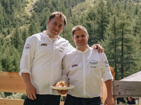 Two chefs are smiling in nature. In the background, green mountains and trees can be seen.