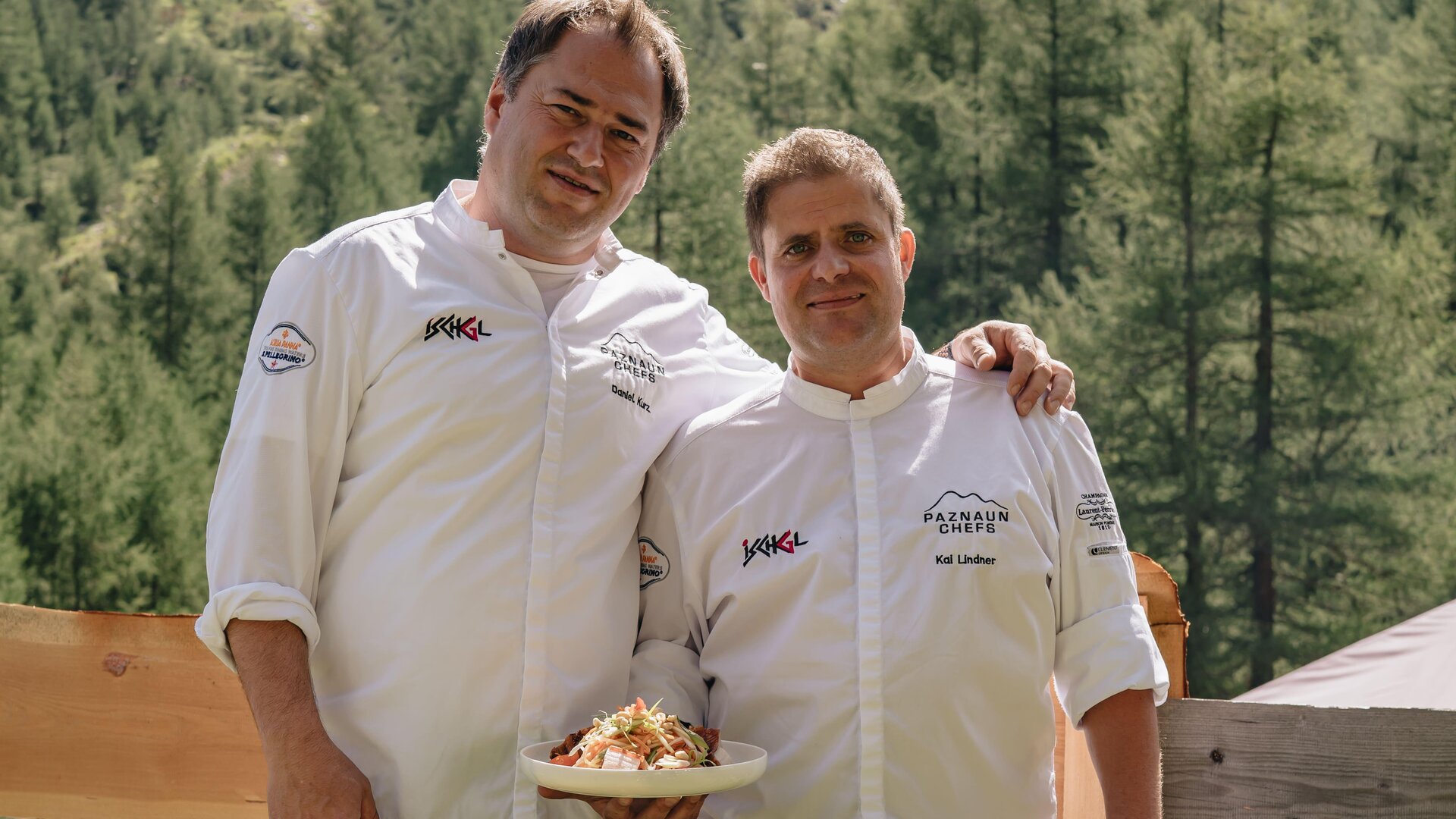 Two chefs are smiling in nature. In the background, green mountains and trees can be seen.
