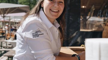 A smiling young woman stands at a wooden table. She is wearing a white shirt and appears friendly and inviting.