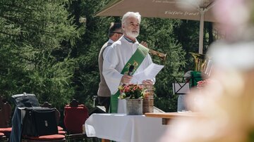 A priest is standing outdoors behind a table with flowers and equipment for a ceremony. In the background, chairs and trees can be seen.