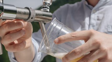 A young man is tapping a drink from a barrel into a plastic cup. He is wearing sunglasses and a hat while standing in a sunny environment.
