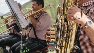 A music band is playing outdoors while wearing traditional costumes. In the foreground, a musician with a tuba can be seen.