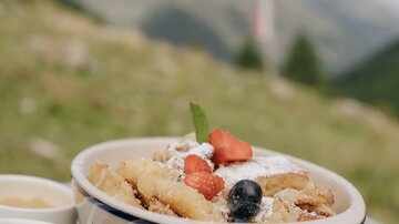 A delicious dish with fried apple pieces, fresh fruits, and powdered sugar. On the side, there are two small bowls of jam and cream.