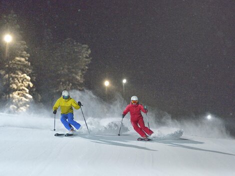 Two skiers are skiing at night on a snow-covered slope. The surroundings are illuminated, creating a winter atmosphere.