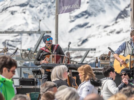 A lively music event in the mountains with snow-capped peaks in the background. Various musicians are performing in front of an audience in the sun.