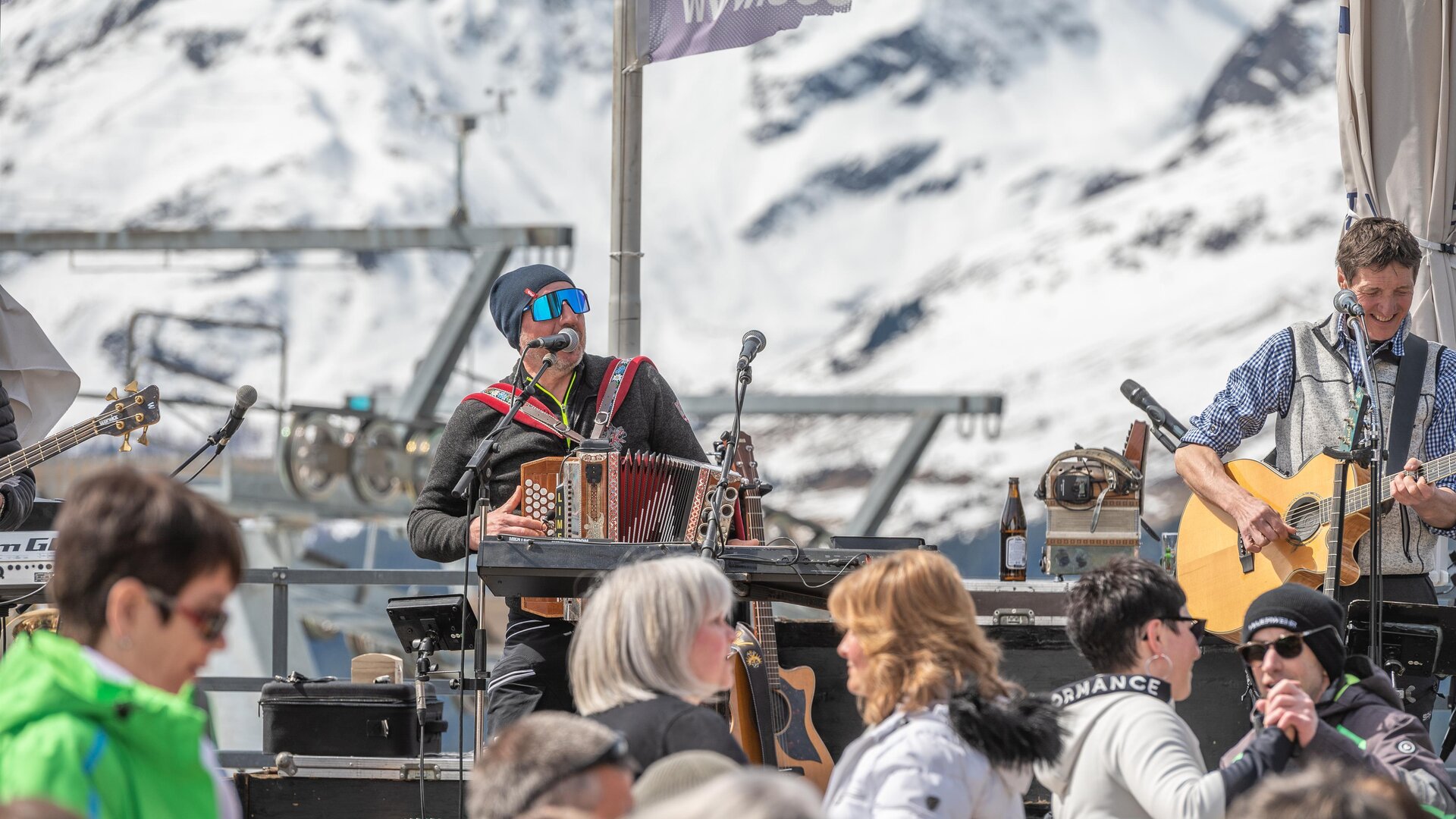 A lively music event in the mountains with snow-capped peaks in the background. Various musicians are performing in front of an audience in the sun.
