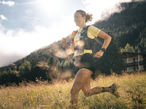 A running person in nature with sunlight in the background. Green meadows and mountains are visible.
