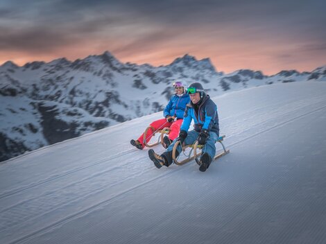 Two people are sitting on sleds and enjoying the snow on a mountain. In the background, there are snow-covered mountains and a beautiful sunset.