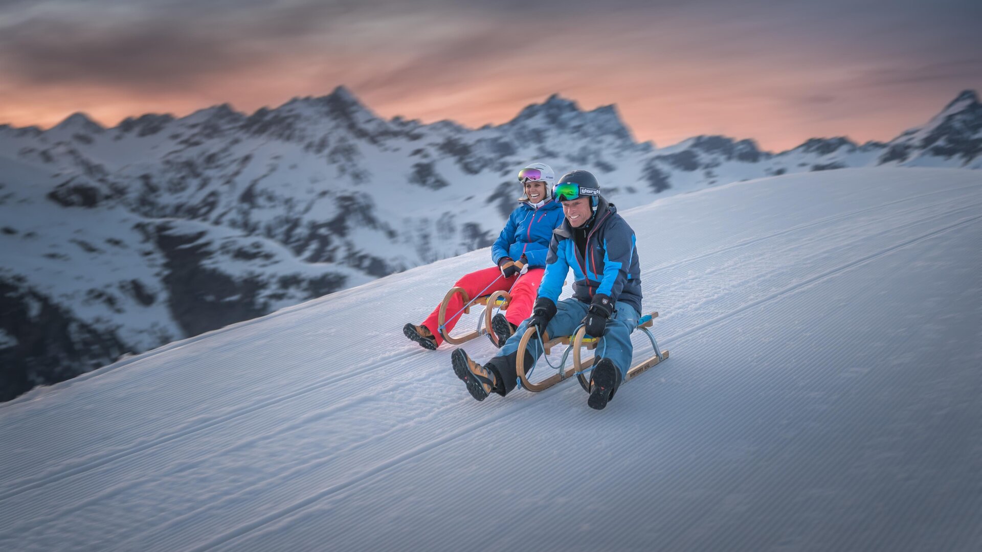 Two people are sitting on sleds and enjoying the snow on a mountain. In the background, there are snow-covered mountains and a beautiful sunset.