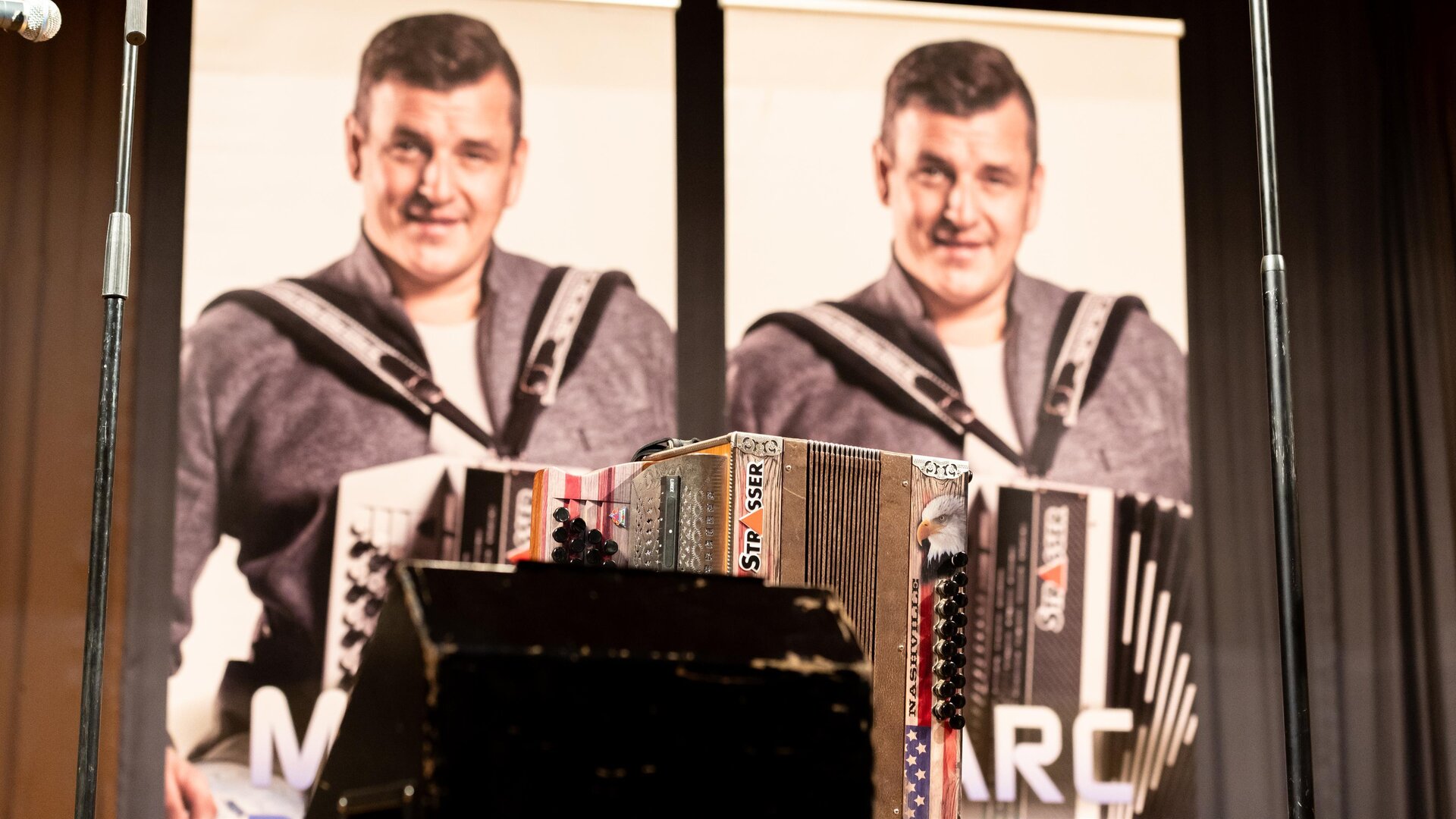 Two posters featuring a smiling man holding an accordion. In the foreground, another accordion is visible.