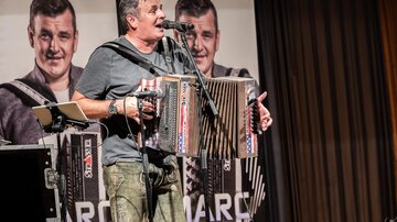 A musician is playing the accordion on a stage. In the background, there are posters featuring his image.