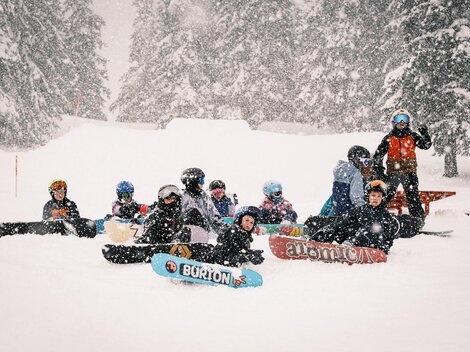A group of snowboarders sits in the snow during a snowstorm. Around them are snow-covered trees and a wintry landscape.