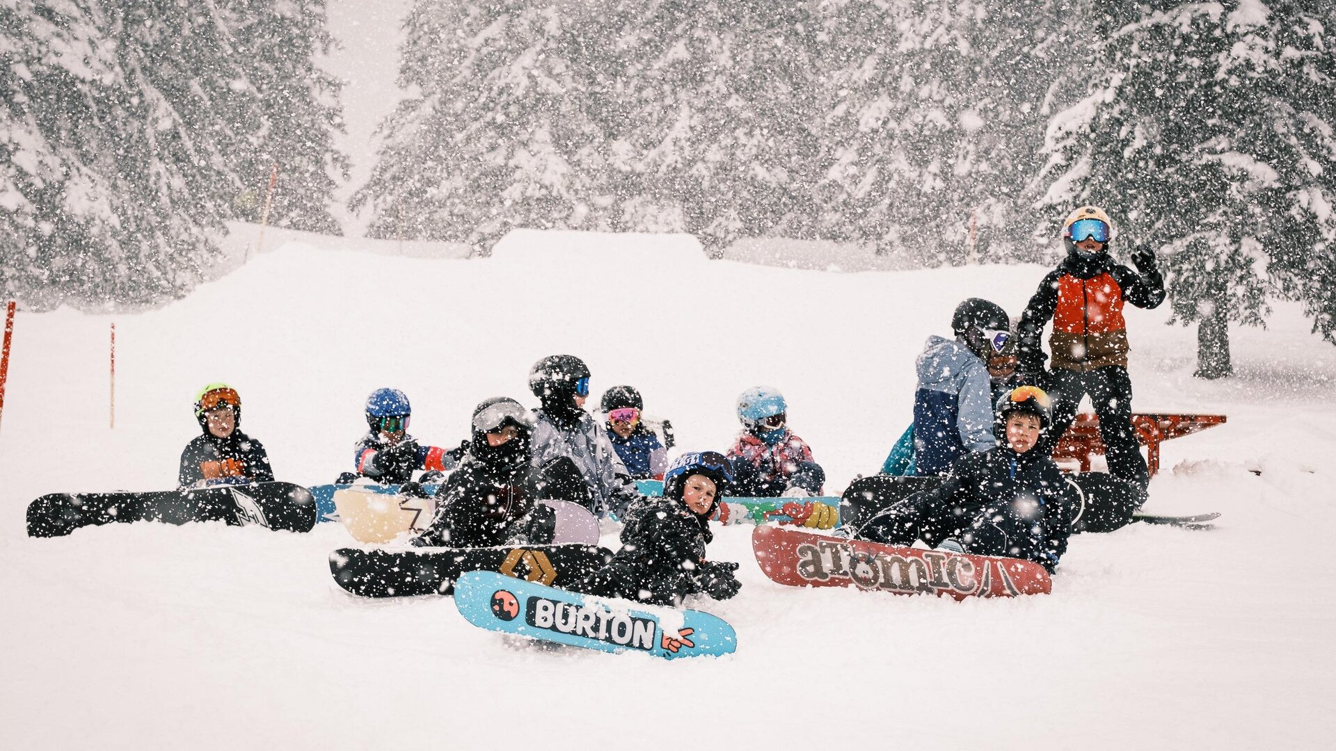 A group of snowboarders sits in the snow during a snowstorm. Around them are snow-covered trees and a wintry landscape.