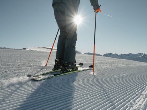 Ein Skifahrer steht auf einer frisch präparierten Piste im Sonnenschein. Im Hintergrund sind schneebedeckte Berge zu sehen.