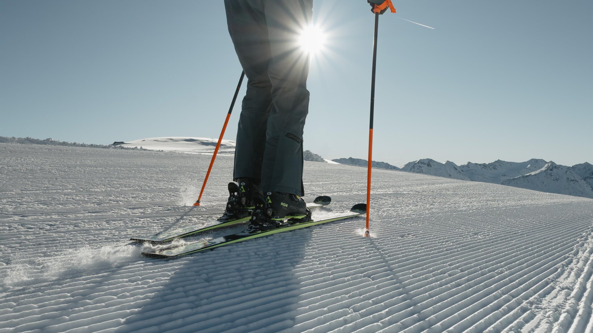 Ein Skifahrer steht auf einer frisch präparierten Piste im Sonnenschein. Im Hintergrund sind schneebedeckte Berge zu sehen.