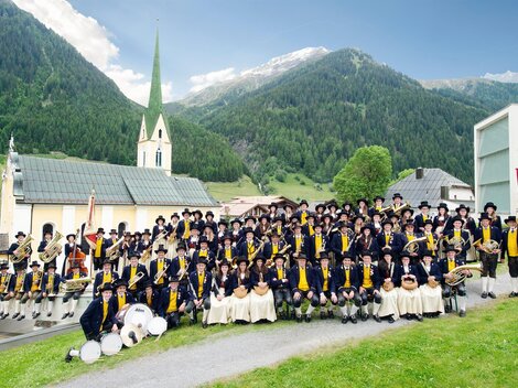 Eine große Gruppe von Menschen in traditionellen Trachten steht in einer malerischen Landschaft. Im Hintergrund sieht man eine Kirche und die Berge der Alpen.