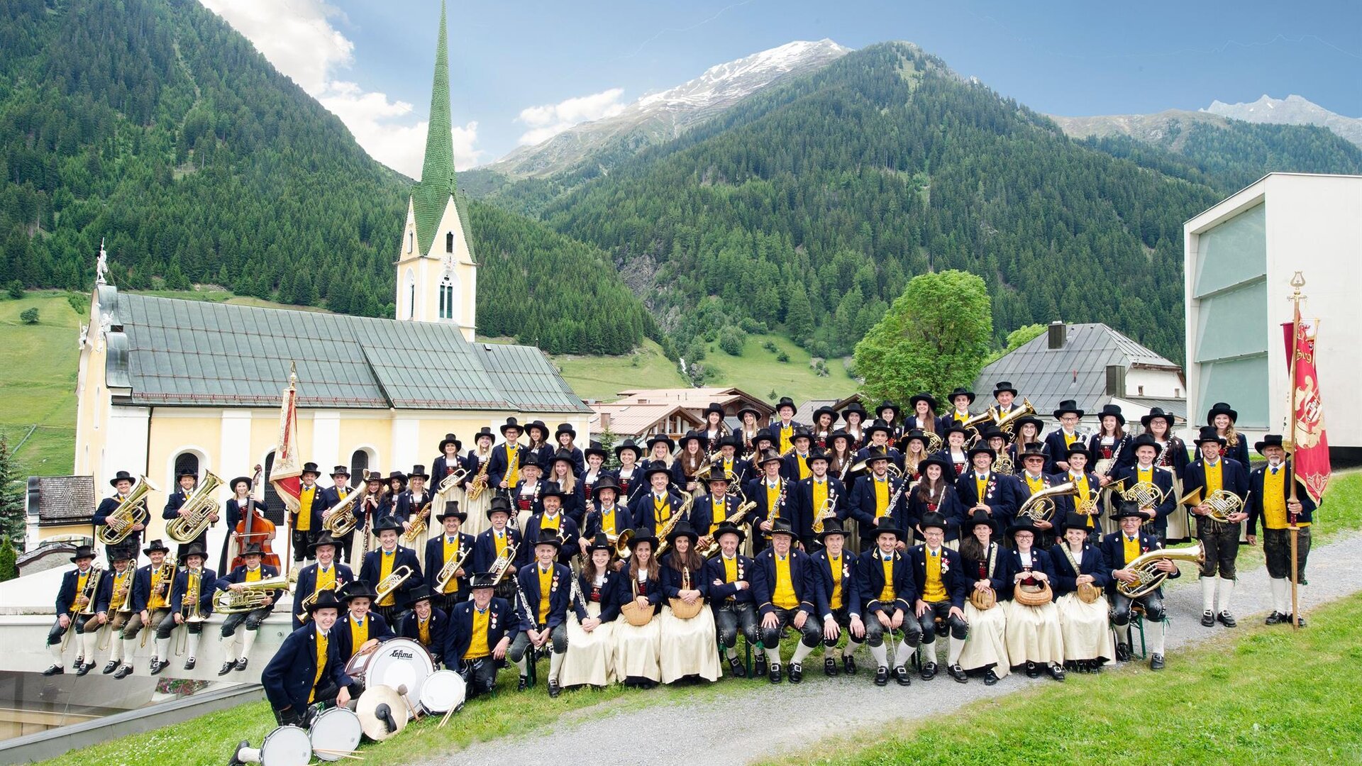 A large group of people in traditional costumes stands in a picturesque landscape. In the background, there is a church and the mountains of the Alps.