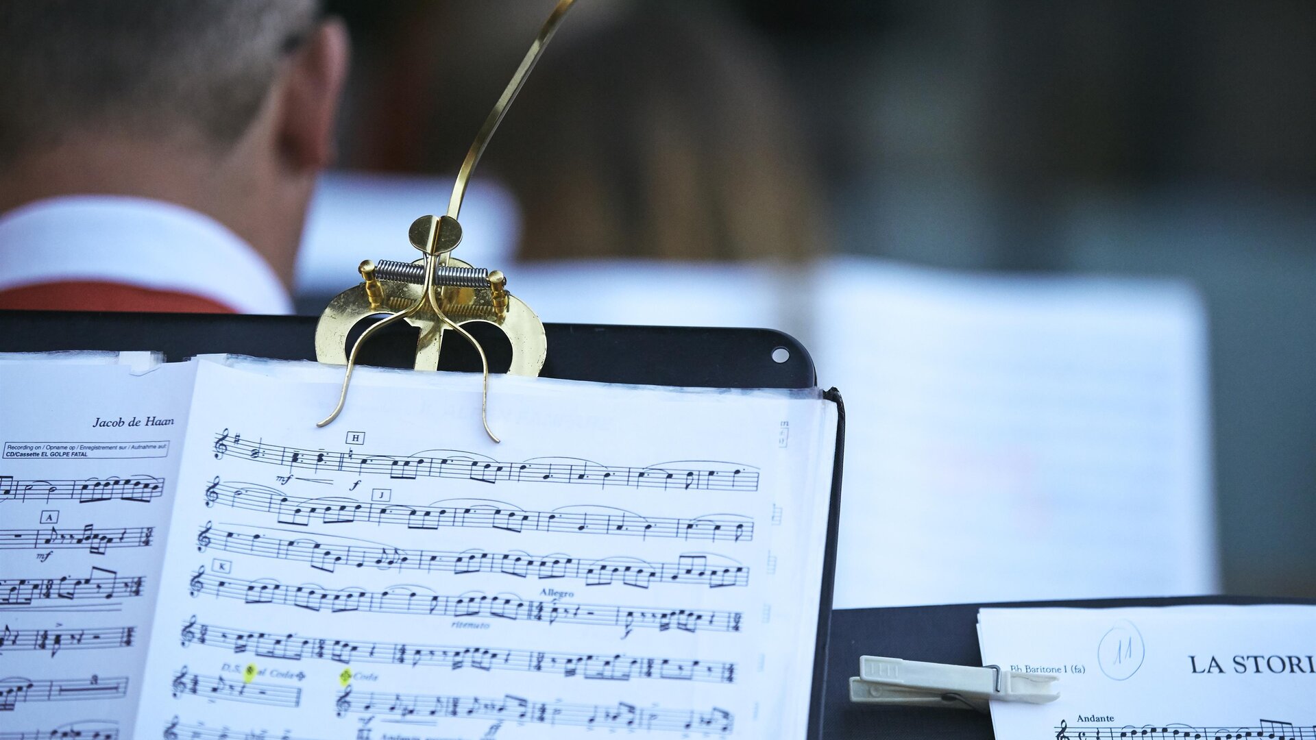 A snippet of sheet music on a music stand. In the background, musicians can be seen preparing for a performance.