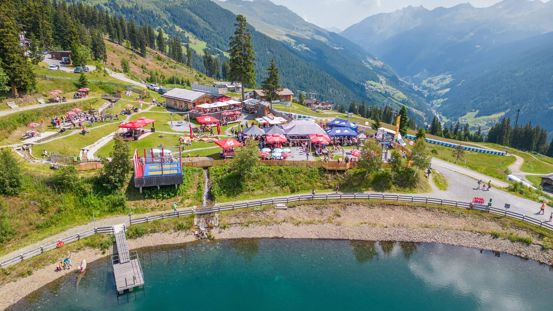A picturesque mountain landscape with a clear lake and numerous colorful umbrellas. In the background, the green slopes of the mountains and a radiant blue sky can be seen.