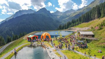 A lively event in the mountains with many people gathering around a clear lake. Colorful tents and a beautiful natural landscape surround the scene.